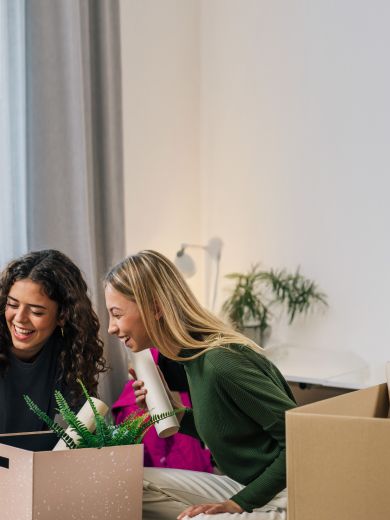 Zwei Frauen sitzen lächelnd auf einem Bett mit Umzugskartons