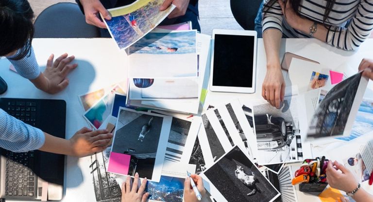 Auf einem Tisch liegen viele Blätter, ein Tablet und ein Laptop. Mehrere Hände sehen beschäftigt aus.