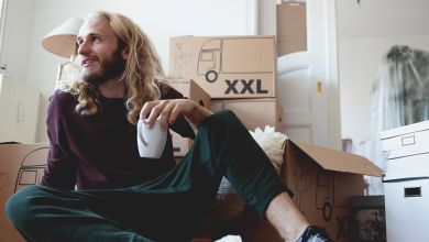 E WIE EINFACH Umzugsservice Junger Mann mit weißer Tasse in der linken Hand. Sitzt auf dem Boden in einer Wohnung vor ein paar gestapelten Umzugskartons.