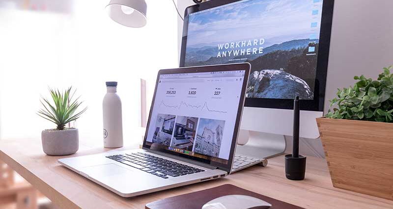 Ein Schreibtisch, auf dem ein aufgeklappter Laptop und ein Computer steht. Auf dem Bildschirm des Computer steht "workhard anywhere". Auf dem Schreibtisch befinden sich auch Pflanzen, eine Wasserflasche und eine Maus zum bedienen der Computer.