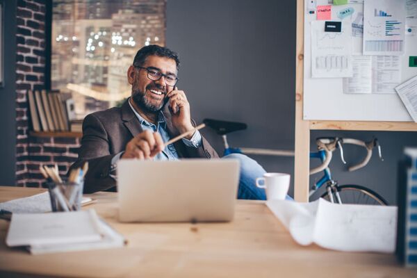 Mann mit Brille sitzt lachend vor einem Laptop am Schreibtisch, hält dabei einen Stift in der linken und ein Handy am Ohr in der rechten Hand.