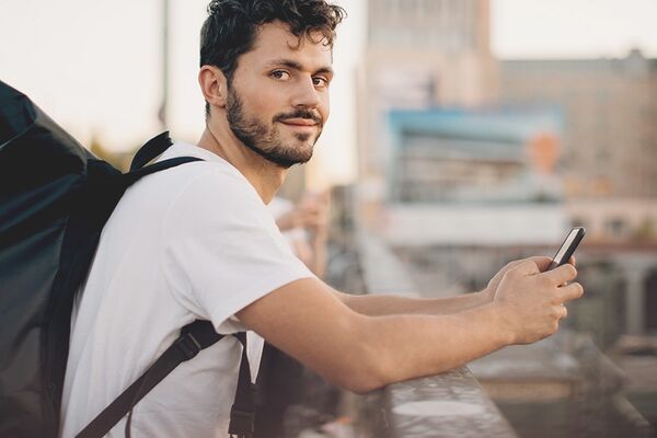 junger Mann mit Rucksack auf dem Rücken und Handy in der Hand schaut freundlich in die Kamera