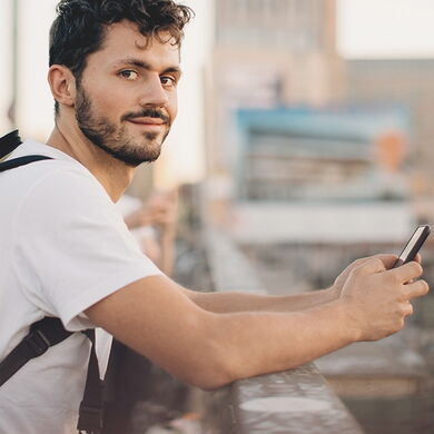 junger Mann mit Rucksack auf dem Rücken und Handy in der Hand schaut freundlich in die Kamera