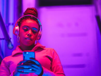Eine Frau steht im pinken Licht mit Kopfhörern auf und schaut auf ihr Handy