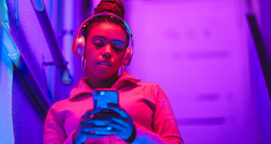 Frau steht im pinken Licht mit Kopfhörern auf und schaut auf ihr Handy