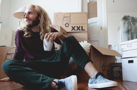 Junger Mann mit weißer Tasse in der linken Hand. Sitzt auf dem Boden in einer Wohnung vor ein paar gestapelten Umzugskartons.
