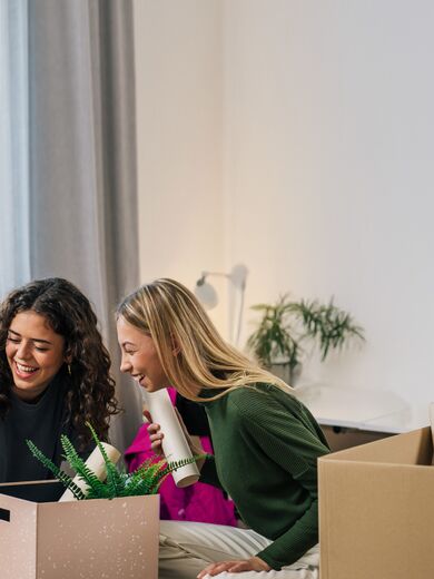 Zwei Frauen sitzen lächelnd auf einem Bett mit Umzugskartons