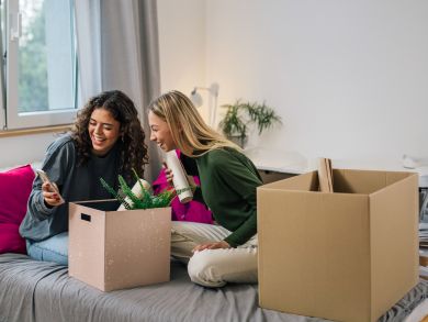Zwei Frauen ziehen um Zwei Frauen sitzen lächelnd auf einem Bett mit Umzugskartons