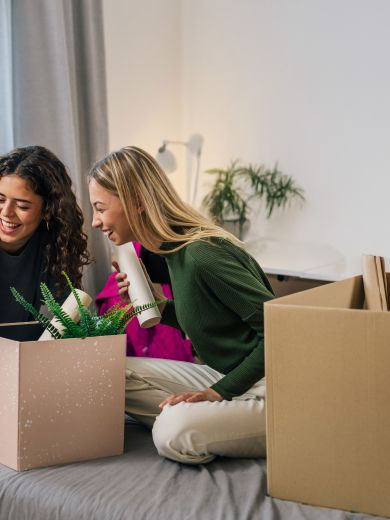 Zwei Frauen sitzen lächelnd auf einem Bett mit Umzugskartons
