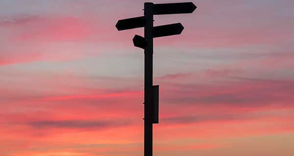 Ein schwarzer Richtungspfeiler, der in verschiedene Richtungen zeigt, vor einem rosafarbenen Himmel.