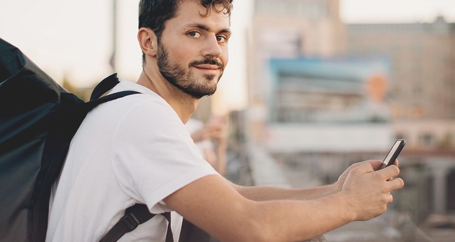 junger Mann mit Rucksack auf dem Rücken und Handy in der Hand schaut freundlich in die Kamera