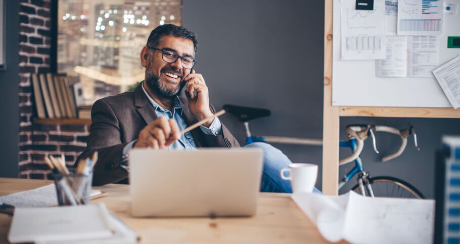 Mann mit Brille sitzt lachend vor einem Laptop am Schreibtisch, hält dabei einen Stift in der linken und ein Handy am Ohr in der rechten Hand.