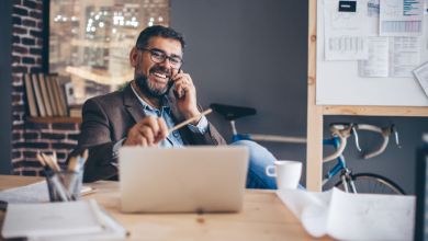 Mann mit Brille sitzt lachend vor einem Laptop am Schreibtisch, hält dabei einen Stift in der linken und ein Handy am Ohr in der rechten Hand.