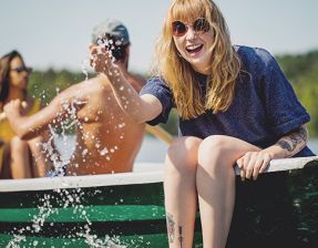 Eine lachende Frau sitzt in einem Boot, mit den Füßen im Wasser und spritzt Wasser in Richtung Kamera. Im Hintergrund zwei weitere Personen, die das Boot steuern.