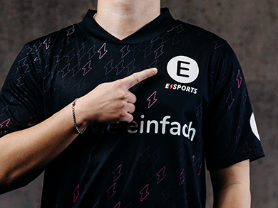 eine person zeigt auf die eigene Brust an die Stelle auf der auf dem Trikot das Logo von E WIE EINFACH E-Sports zu sehen ist