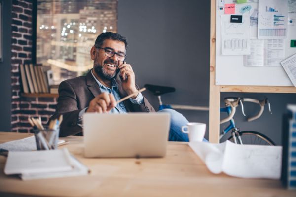 Mann mit Brille sitzt lachend vor einem Laptop am Schreibtisch, hält dabei einen Stift in der linken und ein Handy am Ohr in der rechten Hand.