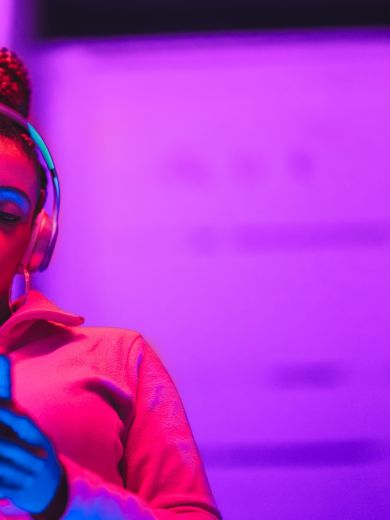 Eine Frau steht im pinken Licht mit Kopfhörern auf und schaut auf ihr Handy