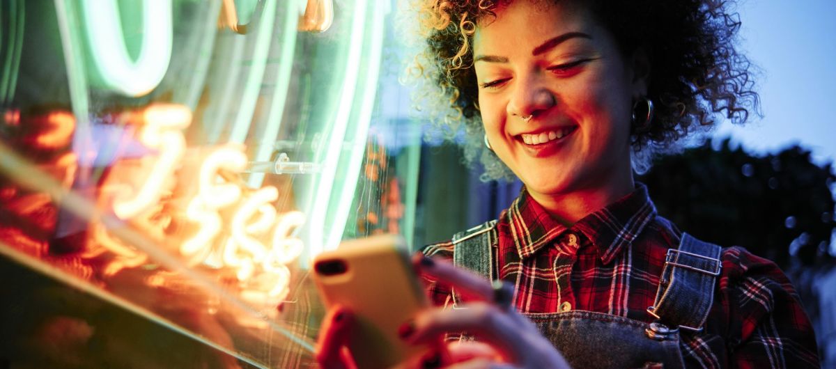 Frau mit lockigen kurzen Haaren steht vor einem Neonlicht und schaut lächelnd auf ihr Handy