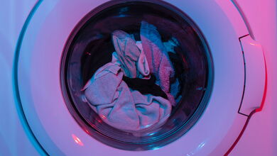 Eine Waschmaschine, die in neonfarbenen Licht angestrahlt wird. Man sieht Wäsche durch ein Fenster an der Tür der Waschmaschine.