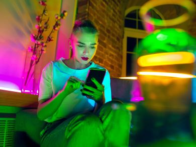 Eine Frau sitzt mit einem Handy auf der Couch und ist umgeben von Neonlichtern
