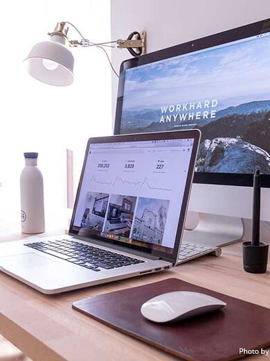 Ein Schreibtisch, auf dem ein aufgeklappter Laptop und ein Computer steht. Auf dem Bildschirm des Computer steht "workhard anywhere". Auf dem Schreibtisch befinden sich auch Pflanzen, eine Wasserflasche und eine Maus zum bedienen der Computer.