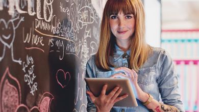 Junge Frau steht mit Tablet in der Hand neben einer bunt bemalten Kreidewand und lächelt dabei freundlich in die Kamera.