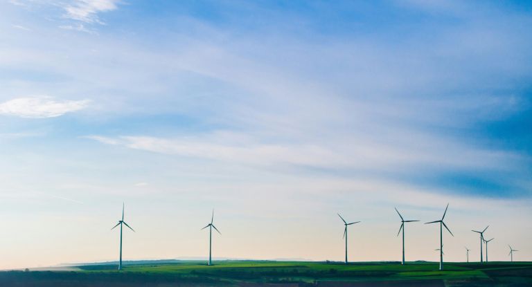 Mehrere Windkraftwerke erzeugen Strom auf einer Wiese vor einem blauen und leicht wolkigem Himmel