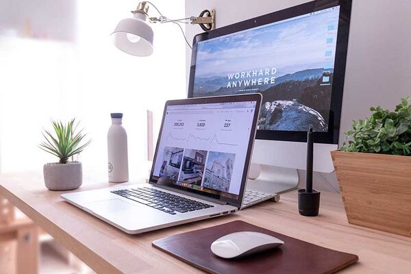 Ein Schreibtisch, auf dem ein aufgeklappter Laptop und ein Computer steht. Auf dem Bildschirm des Computer steht "workhard anywhere". Auf dem Schreibtisch befinden sich auch Pflanzen, eine Wasserflasche und eine Maus zum bedienen der Computer.