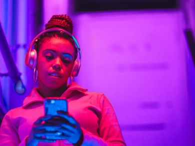 Eine Frau steht im pinken Licht mit Kopfhörern auf und schaut auf ihr Handy