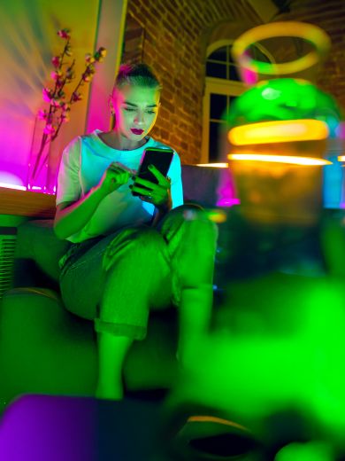 Eine Frau sitzt mit einem Handy auf der Couch und ist umgeben von Neonlichtern