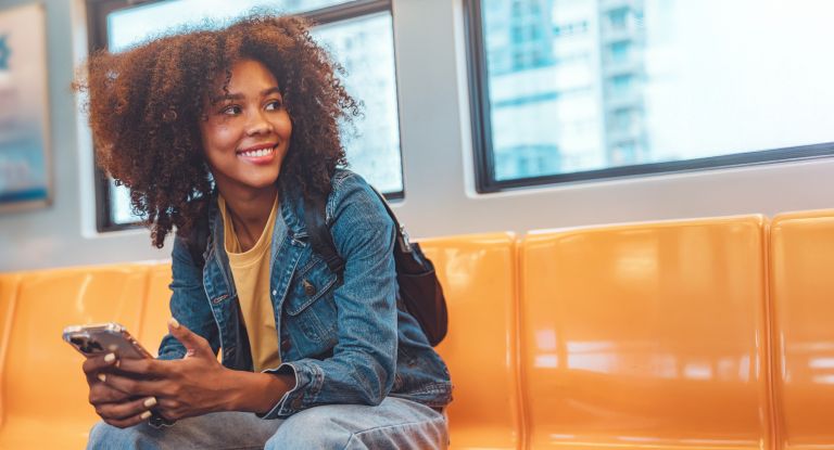 Eine junge, glückliche Frau die in einem leeren Zug sitzt und ein Smartphone in der Hand hält.