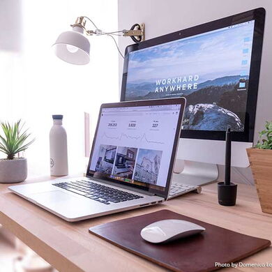 Ein Schreibtisch, auf dem ein aufgeklappter Laptop und ein Computer steht. Auf dem Bildschirm des Computer steht "workhard anywhere". Auf dem Schreibtisch befinden sich auch Pflanzen, eine Wasserflasche und eine Maus zum bedienen der Computer.