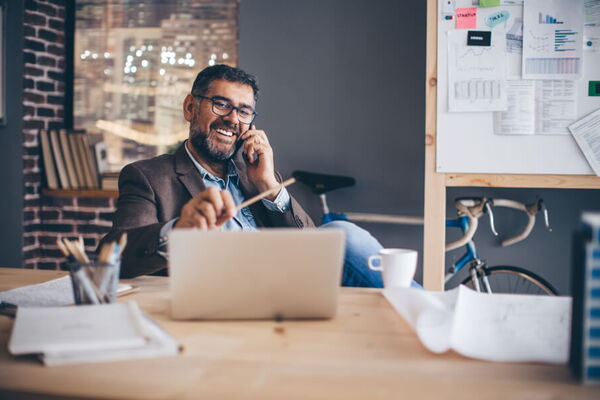 Mann mit Brille sitzt lachend vor einem Laptop am Schreibtisch, hält dabei einen Stift in der linken und ein Handy am Ohr in der rechten Hand.