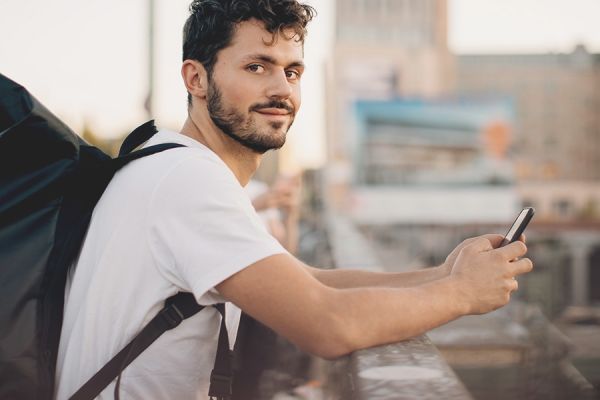 junger Mann mit Rucksack auf dem Rücken und Handy in der Hand schaut freundlich in die Kamera