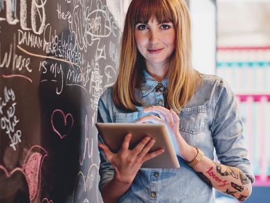 Junge Frau steht mit Tablet in der Hand neben einer bunt bemalten Kreidewand und lächelt dabei freundlich in die Kamera.